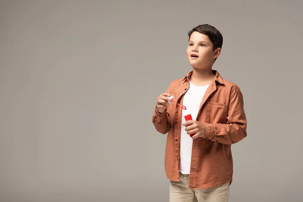 Amazed boy holding bottle with soap bubbles and looking away isolated on grey — Stock Photo