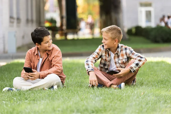Два улыбающихся брата разговаривают, сидя на газоне со смартфонами — стоковое фото