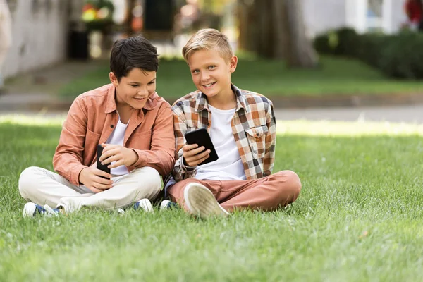 Два улыбающихся брата сидят на зеленой траве и используют смартфоны — Stock Photo