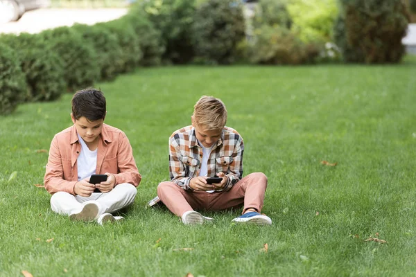 Deux frères concentrés assis sur la pelouse verte et utilisant des smartphones — Photo de stock