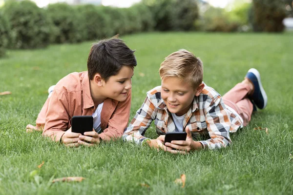 Two cheerful brothers lying on green lawn and using smartphones — Stock Photo