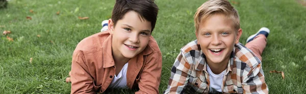 Panoramic shot of two cheerful brothers lying on green grass and smiling at camera — Stock Photo