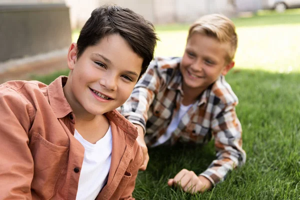 Selektiver Fokus des fröhlichen Jungen, der in die Kamera blickt, während er in der Nähe seines Bruders auf dem Gras liegt — Stockfoto