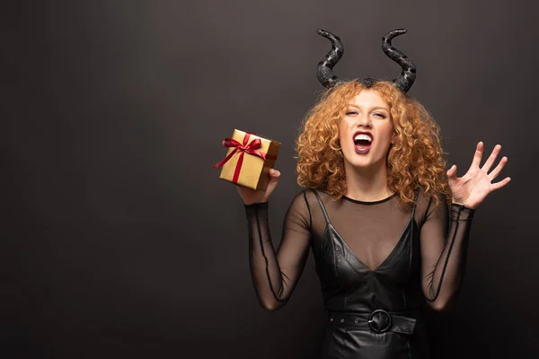 Beautiful redhead woman in halloween costume with horns holding gift box on black — Stock Photo