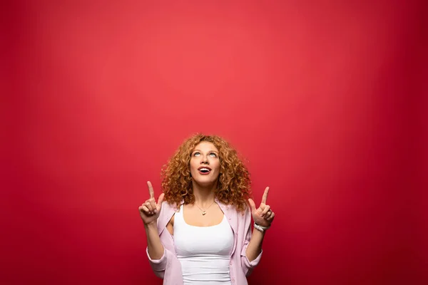 Hermosa mujer pelirroja sorprendida señalando hacia arriba, aislado en rojo - foto de stock