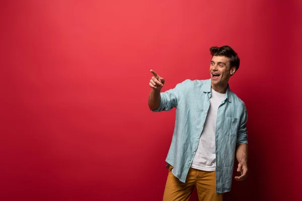 Handsome laughing man in casual clothes pointing isolated on red — Stock Photo