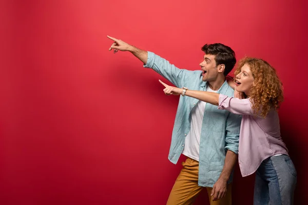 Beau couple excité en vêtements décontractés pointant isolé sur rouge — Photo de stock