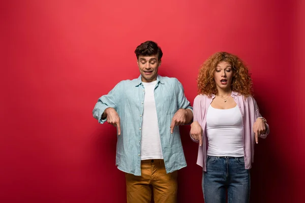 Belo casal chocado em roupas casuais apontando para baixo, isolado no vermelho — Fotografia de Stock