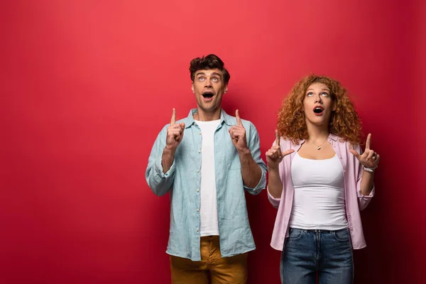 Hermosa pareja sorprendida en ropa casual apuntando hacia arriba, aislado en rojo - foto de stock