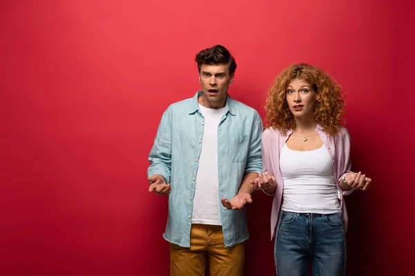 Beautiful confused couple gesturing in casual clothes isolated on red — Stock Photo