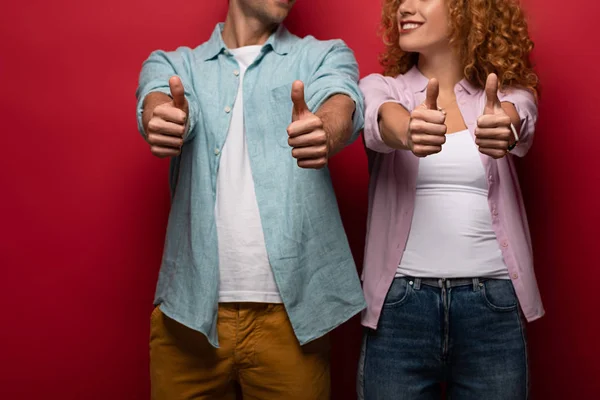 Vista cortada de casal sorrindo mostrando polegares para cima, isolado em vermelho — Fotografia de Stock