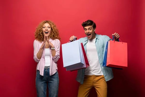 Excitada pareja joven sosteniendo bolsas de compras, aislado en rojo - foto de stock