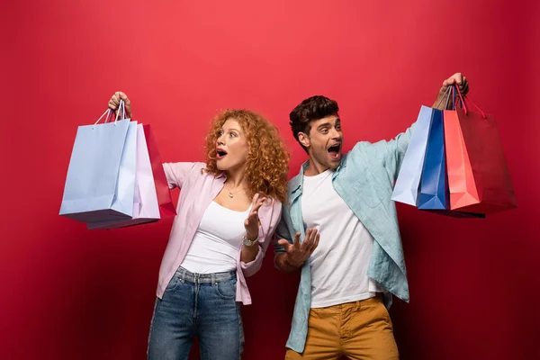 Chocado casal olhando para sacos de compras, isolado no vermelho — Fotografia de Stock