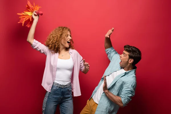 Attractive curly woman throwing little rocket on scared man, on red — Stock Photo