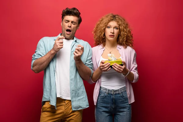 Sick man sneezing while sad woman holding paper napkins, on red — Stock Photo