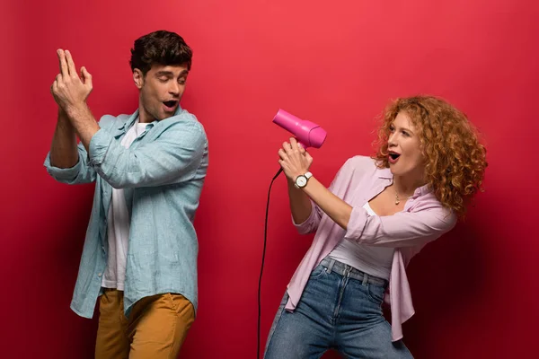 Young couple having fun with hair dryer and hand gun, on red — Stock Photo