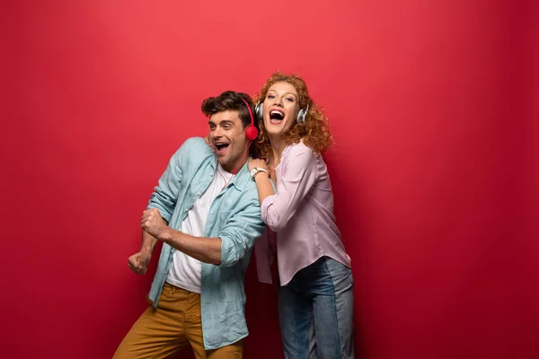 Pareja emocionada bailando y escuchando música con auriculares, aislados en rojo - foto de stock