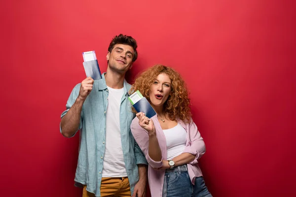 Pareja de viajeros sonrientes sosteniendo pasaportes con billetes de avión, aislados en rojo - foto de stock