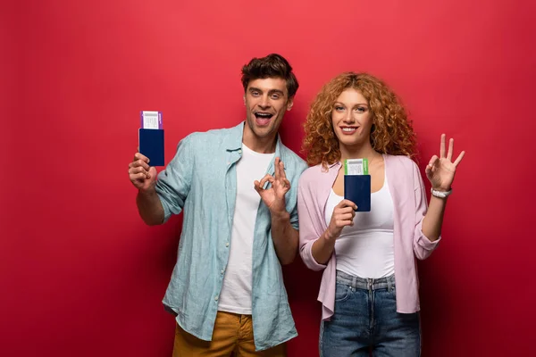 Par de viajantes felizes segurando passaportes com bilhetes aéreos enquanto mostra sinal ok, isolado no vermelho — Fotografia de Stock