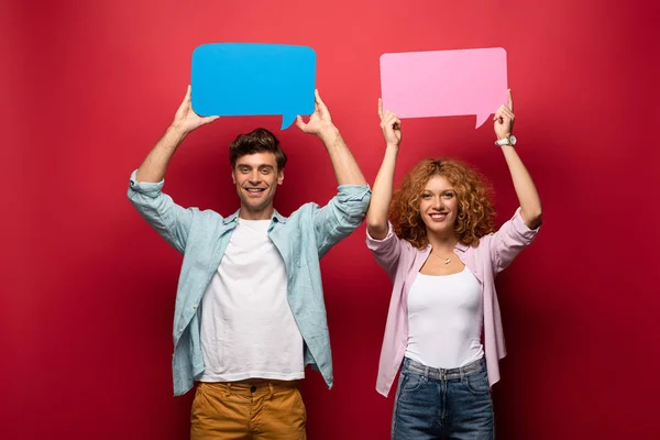 Sonriente pareja sosteniendo burbujas de habla rosa y azul, en rojo - foto de stock