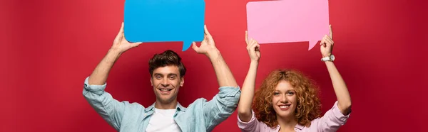 Cheerful couple holding pink and blue speech bubbles, on red — Stock Photo