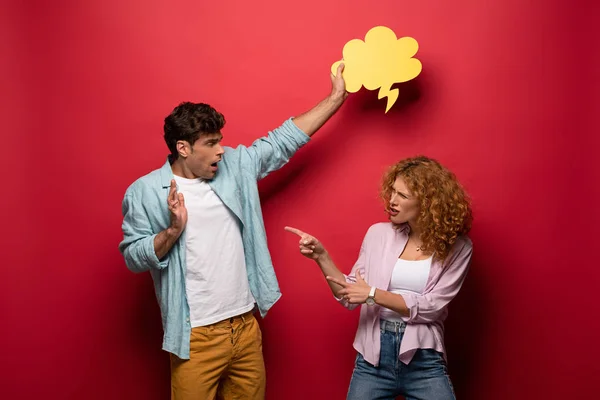 Boyfriend holding yellow cloud bubble over angry girlfriend, on red — Stock Photo