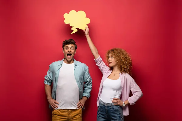 Smiling girlfriend holding yellow cloud bubble over boyfriend, on red — Stock Photo