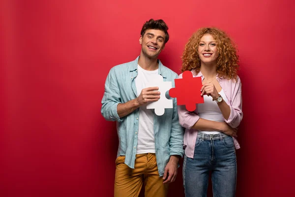 Cheerful couple holding puzzle pieces, isolated on red — Stock Photo