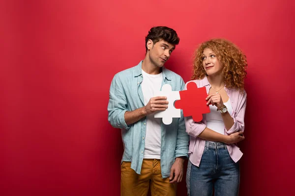 Sorrindo casal segurando peças de quebra-cabeça e olhando um para o outro, isolado em vermelho — Fotografia de Stock