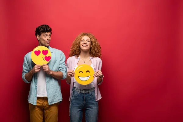 KYIV, UKRAINE - SEPTEMBER 26, 2019: cheerful couple holding smiling and kissing emoji signs, on red — Stock Photo