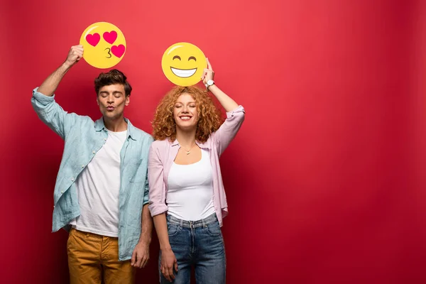KYIV, UCRANIA - 26 de septiembre de 2019: pareja feliz sosteniendo carteles emojis sonrientes y besándose, en rojo - foto de stock