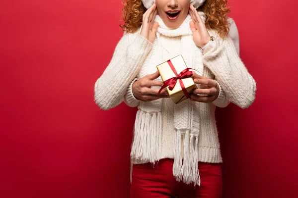 Vista recortada de la mujer sorprendida y el hombre en traje de invierno celebración de regalo de Navidad aislado en rojo - foto de stock