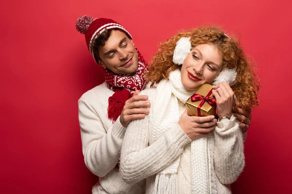 Boyfriend and girlfriend in winter outfit holding christmas gift isolated on red — Stock Photo