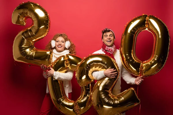 Schönes Paar im Winteroutfit mit goldenen Luftballons für Neujahr 2020, auf rot — Stockfoto