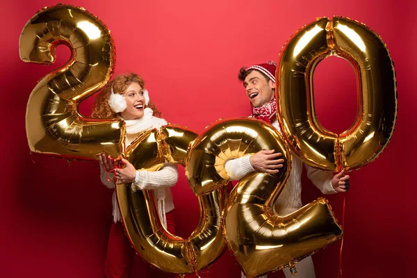 Улыбающаяся пара в зимнем наряде, держащая золотые воздушные шары нового 2020 года, на красном — стоковое фото