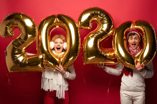 Excited couple in winter outfit holding new year 2020 golden balloons, on red — Stock Photo