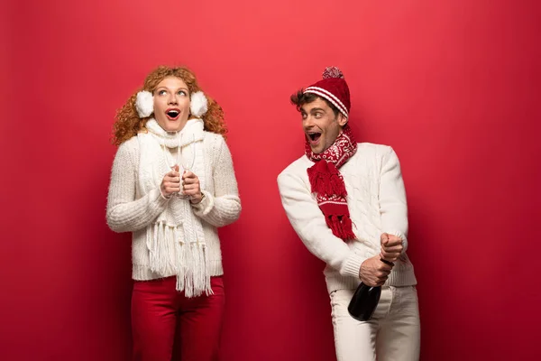 Couple excité en tenue d'hiver tenant champagne pour célébrer Noël, sur rouge — Photo de stock