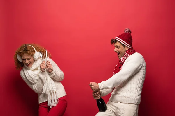 Cheerful funny couple in winter outfit holding champagne for celebrating christmas, on red — Stock Photo