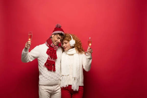 Beautiful couple in winter outfit hugging and holding champagne glasses, on red — Stock Photo