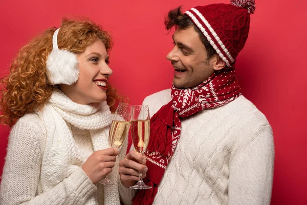 Beautiful smiling couple in winter outfit clinking with champagne glasses, isolated on red — Stock Photo