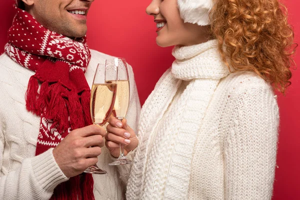 Vista recortada de la pareja sonriente en traje de invierno tintineo con copas de champán, aislado en rojo - foto de stock
