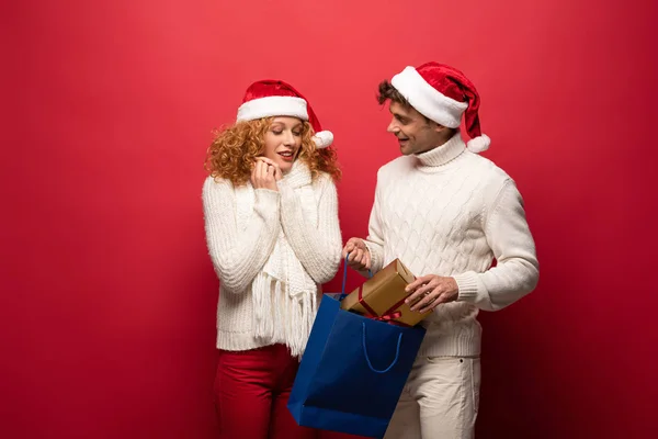 Beau couple dans chapeaux Santa avec présent dans le sac à provisions, isolé sur rouge — Photo de stock