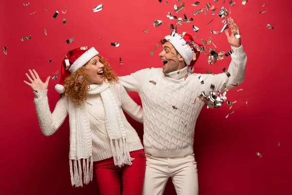 Couple excité dans chapeaux de Père Noël célébrant avec des confettis dorés, isolé sur rouge — Photo de stock