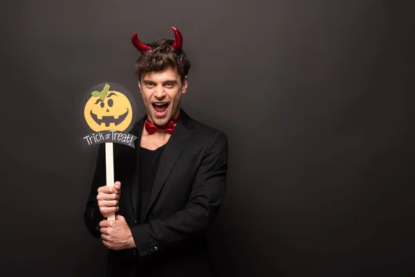 Sexy man in devil costume holding pumpkin trick or treat sign on black — Stock Photo