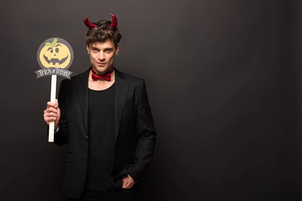 Handsome man in devil costume holding pumpkin trick or treat sign on black — Stock Photo