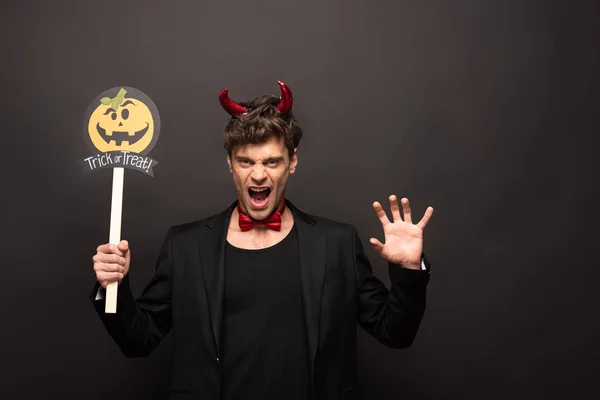 Handsome sexy man in halloween devil costume holding pumpkin trick or treat sign on black — Stock Photo