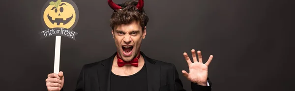 Brunette man in devil costume holding pumpkin trick or treat halloween sign on black — Stock Photo