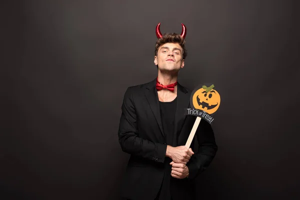 Handsome young man in halloween devil costume holding pumpkin trick or treat sign on black — Stock Photo
