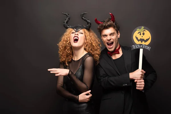 Scary couple posing in halloween costumes with pumpkin trick or treat sign on black — Stock Photo