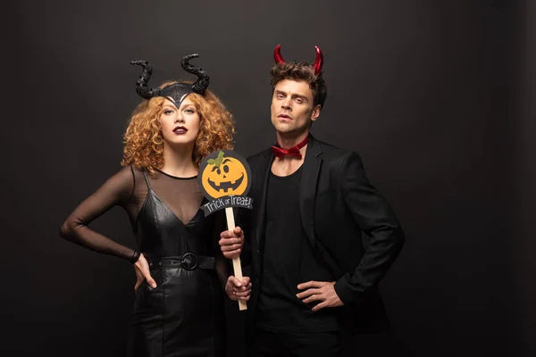 Beau couple posant en costumes d'Halloween avec tour de citrouille ou traiter signe sur noir — Photo de stock